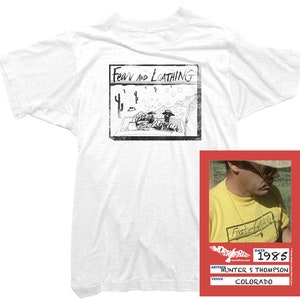 Puede incluir: Camiseta blanca con una ilustraci&oacute;n en blanco y negro de un coche conduciendo por un paisaje des&eacute;rtico. La ilustraci&oacute;n se titula "Fear and Loathing". La camiseta es de estilo vintage, con un aspecto desgastado. La imagen tambi&eacute;n incluye un primer plano de un hombre que lleva una camiseta amarilla con la misma ilustraci&oacute;n. La camiseta est&aacute; fechada en 1985 y dice "Hunter S. Thompson, Colorado".