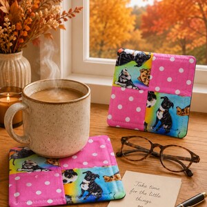 May include: Two square fabric coasters featuring a patchwork design of pink fabric with white polka dots and a colourful print of various dog breeds. A speckled ceramic mug with hot coffee, brown-rimmed glasses, and a note with the text "Take time for the little things" are also present.