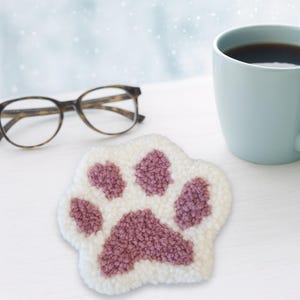 May include: A white and mauve paw print coaster with a looped texture. The coaster is on a white surface, next to a pair of spectacles and a light blue mug filled with coffee.