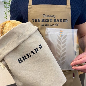 Może przedstawiać: Osoba w beżowym fartuchu z napisem "Probably THE BEST BAKER in the world" trzyma bochenek chleba, torbę na chleb z napisem "BREAD" i przezroczystą plastikową torbę ze wzorem pszenicy.