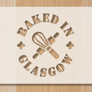 May include: A light-coloured wooden sign featuring the text "BAKED IN GLASGOW" in a circular arrangement. A whisk and rolling pin are at the centre. Four floral cut-out decorations are also present.
