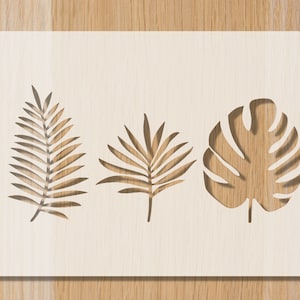 Peut inclure: Un panneau en bois blanc avec trois motifs de feuilles tropicales découpés. Les feuilles sont disposées en rangée, avec une feuille de palmier à gauche, une feuille de palmier au centre et une feuille de monstera à droite.