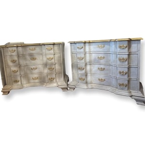 May include: Two antique dressers with a light gray finish and gold accents. Each dresser has four drawers with ornate gold handles and a curved front. The top edges are trimmed with gold. The dressers are on a white background.