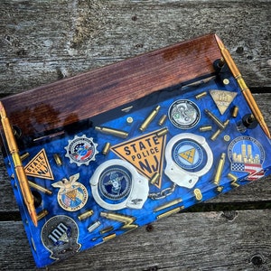 May include: A rectangular serving tray featuring a dark wood top section and a blue resin base embedded with various New Jersey State Police memorabilia, including metal badges, handcuffs, and brass bullet casings, with two gold-toned bullet handles.
