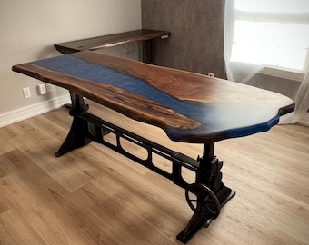 May include: A large wooden table with a blue epoxy river running through the center. The table is supported by a black metal base with a wheel.