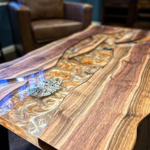 Peut inclure: Une table basse en bois avec un design à bord vif et un plateau en résine époxy transparente. La résine présente un motif tourbillonnant et marbré dans des tons d'or et de blanc. Un petit modèle argenté du vaisseau spatial Millennium Falcon est intégré à la résine.