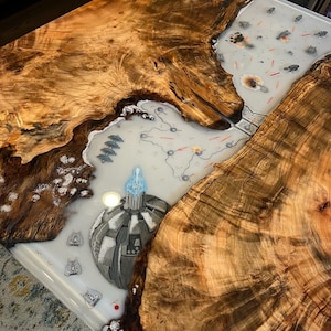 Peut inclure: Un plateau de table en bois avec un incrustation de résine époxy transparente représentant une scène de bataille spatiale détaillée. La scène comprend des vaisseaux spatiaux, des explosions et une grande structure ronde grise avec une flamme bleue.