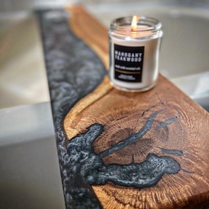 May include: A wooden tray with a black and silver epoxy resin inlay. The tray has a natural wood grain and a knot in the center. A candle with a white label that reads "MANOGANY TEAKWOOD" is sitting on the tray.