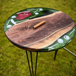 May include: A round wooden table with a green and brown epoxy resin top featuring a golf course design with golf balls, tees, and a flag. The table has three black hairpin legs.