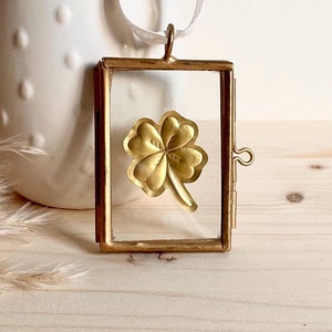 Puede incluir: Un amuleto de trébol de cuatro hojas dorado dentro de un marco de vidrio rectangular. El marco tiene un cierre de bisagra y está suspendido por una cinta blanca. El fondo incluye un jarrón blanco y una superficie de madera clara.