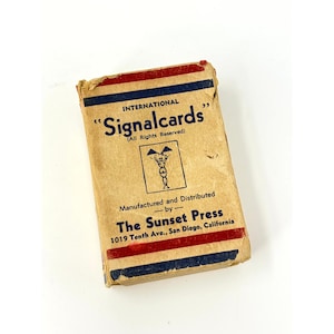 May include: A rectangular tan cardboard box for International Signalcards with blue and red horizontal stripes. The text reads International Signalcards, All Rights Reserved, Manufactured and Distributed by The Sunset Press, 1019 Tenth Ave., San Diego, California.