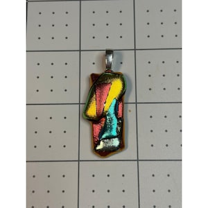 May include: A dichroic glass pendant with a silver bail. The pendant features a rectangular shape with a colorful design of red, yellow, and teal glass. The glass has an iridescent quality, reflecting different colors depending on the light.