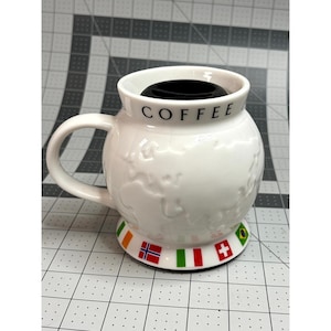May include: White ceramic coffee mug with a world map design and the word "COFFEE" in black. The mug has a handle and a base decorated with flags. The mug is filled with black coffee.