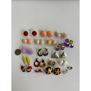 May include: A collection of assorted stud and dangle earrings in various shapes, colors, and materials, including circular, triangular, and floral designs in red, white, peach, yellow, purple, and metallic tones, displayed alongside a US coin.