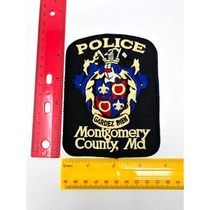 May include: A black embroidered police patch featuring a central crest with red and blue sections, gold accents, and the text POLICE, GARDEZ BIEN, and Montgomery County, Md. A red ruler shows the patch is approximately 4.5 inches tall.