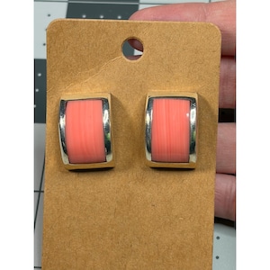 May include: A pair of rectangular clip-on earrings. Each earring has a silver-colored frame with a coral-colored center. The earrings are displayed on a brown cardboard backing.
