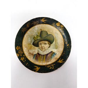 May include: A round decorative plate featuring a central portrait of a person wearing a wide-brimmed hat and a ruffled collar. The border is dark green with gold-colored illustrations of grapevines, leaves, and flying birds on a textured background.