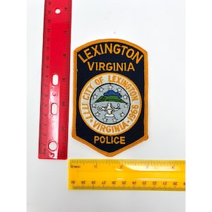 May include: A dark blue embroidered police patch for Lexington, Virginia, featuring a central circular emblem with a lamp and mountain graphic, surrounded by stars and text. A red ruler shows the patch is approximately 6.25 inches tall.