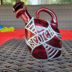 May include: A red glass decanter with a silver metal design and the word "SCOTCH" embossed on the side. The decanter has a stopper with a unique shape.