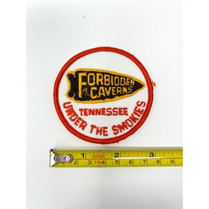May include: A circular embroidered patch with a white background and a red border. It features a yellow and black arrowhead shape with the text "FORBIDDEN CAVERNS" and "TENNESSEE UNDER THE SMOKIES" in red. A yellow measuring tape shows a 3-inch diameter.