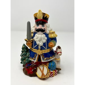 May include: A decorative ceramic nutcracker figurine in a blue uniform with gold accents, holding a sword. The figurine is adorned with a black mustache, white beard, and a tall hat. It is surrounded by a Christmas tree, a toy horse, a drum, and wrapped gifts.