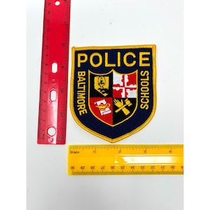 May include: A shield-shaped embroidered patch with a dark blue background and yellow border. It features the text POLICE BALTIMORE SCHOOLS in yellow. The center displays a crest with four quadrants containing a lamp, cross, book, and hand with gavel.