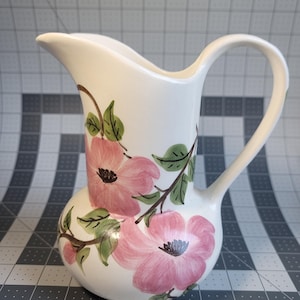 May include: White ceramic pitcher with a curved handle and spout. The pitcher is decorated with pink floral designs and green leaves. The flowers have dark pink centers. The pitcher is a decorative item.