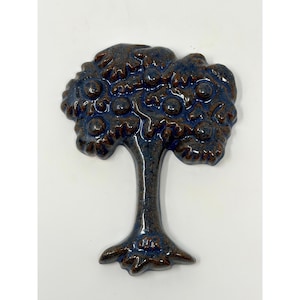 May include: A ceramic wall ornament shaped like a tree with a glossy finish. The piece features a dark brown trunk and roots, while the rounded canopy is a deep blue color with raised circular details representing fruit or leaves throughout.