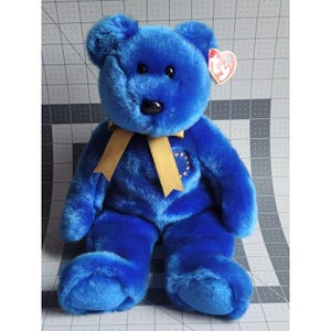 May include: A plush blue teddy bear with a gold ribbon bow and a patch featuring the European Union flag. The bear has a black nose and a Ty heart-shaped tag attached to its ear. The bear is approximately 12 inches tall.