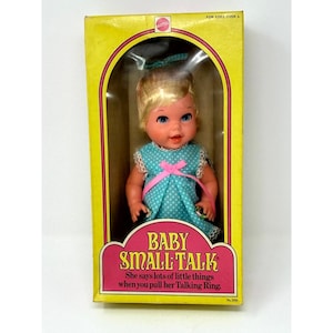 May include: A Mattel Baby Small Talk doll with blonde hair and blue eyes, wearing a teal dress with white polka dots and a pink ribbon. The doll is inside a yellow rectangular box with the text Baby Small Talk and For Ages Over 3 printed.