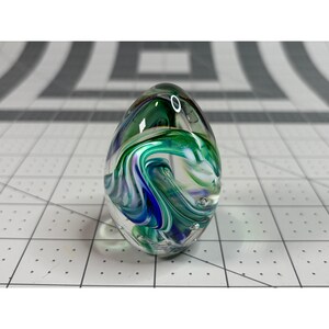 May include: A clear glass paperweight with an egg shape. Swirls of green, blue, and white colors are visible inside the glass. The paperweight has a smooth, rounded surface and is displayed on a grid.