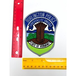 May include: A shield-shaped Burlington Police patch featuring a brown tower, green landscape, and blue sky. The text reads BURLINGTON POLICE, LIBERTAS ET UNITAS, and STATE OF VERMONT. A red ruler shows the patch is approximately 4.5 inches tall.