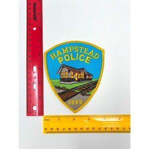 May include: A shield-shaped embroidered patch featuring a blue background, yellow border, and the text "HAMPSTEAD POLICE" above a building illustration and the year "1888". A red ruler shows the patch is approximately 3.5 inches tall.