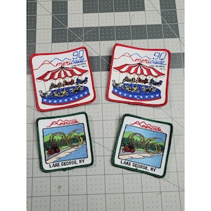 May include: Four embroidered patches. Two patches have a red and white carousel design with the text "90 Americade Lake George N.Y." The other two patches have a green border with a motorcycle and the text "Lake George, NY."