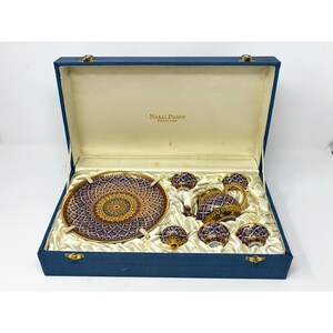 May include: A tea set in a blue fabric-covered presentation box with the text Narai Phand Thailand. The set includes a round plate, a teapot, and four small cups, all featuring a detailed blue, gold, and green geometric pattern.