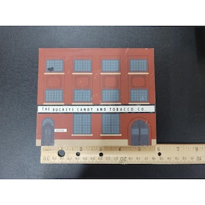 May include: A wooden block featuring a vintage illustration of a brick building with multiple windows and doors. The text "THE BUCKEYE CANDY AND TOBACCO CO." is displayed across the top. The block measures approximately 5 inches in length.