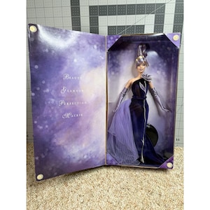 Puede incluir: Una muñeca Barbie en una caja. La muñeca lleva un vestido azul oscuro de un solo hombro con una cola de tul lavanda y guantes plateados. La caja es morada con las palabras "Beauty, Glamour, Perfection, Mackie."