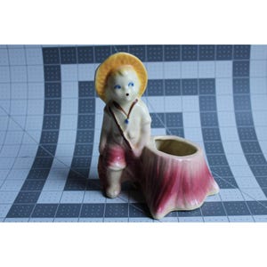 May include: A vintage ceramic planter featuring a young boy in a straw hat and red shorts, leaning against a tree stump-shaped planter. The boy has blue eyes and a small blue pendant. The planter is painted in shades of cream, red, and yellow.