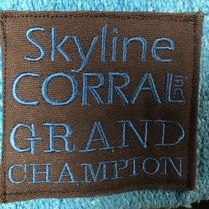 May include: A brown fabric patch with blue embroidered text that reads "Skyline CORRAL GRAND CHAMPION". The patch is rectangular with a stitched border and is set against a light blue textured background.