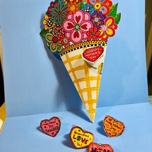 Könnte beinhalten: Ein bunter Blumenstrauß in einem gelben und weißen Kegel mit einem roten Herz-Etikett, auf dem "Happy Mother's Day" steht. Es gibt vier kleinere herzförmige Etiketten mit verschiedenen Botschaften: "Happy Mother's Day Teacher", "Grandma Love Ya!", "Happy Mother's Day with Love" und "Mom Happy Mother's Day".