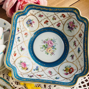 Może przedstawiać: Kwadratowa porcelanowa miska z falistymi brzegami, posiadająca białą podstawę ze złotym wzorem kratki, żywą turkusową obwódkę oraz ręcznie malowane bukiety kwiatów w centrum i w polach kratki, ozdobiona złotymi akcentami.