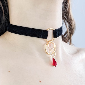 Könnte beinhalten: Ein schwarzer Samt-Choker mit einer goldenen Kette und einem roten Kristall-Anhänger.