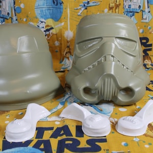 May include: Two unpainted plastic Star Wars Stormtrooper helmets, one with a full face and one with a dome-shaped head, and four white plastic pieces for the helmet's ears.