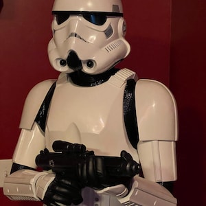 May include: A white Stormtrooper figure from Star Wars, holding a black blaster. The figure has a white helmet with black accents and a black visor. The armor is white with black detailing. The figure is a bust, showing the upper body.
