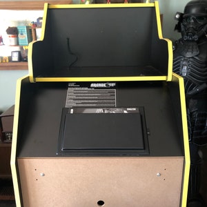 May include: An Arcade1Up cabinet in the process of being assembled. The cabinet has a black interior with yellow trim and the Arcade1Up logo. The cabinet is made of wood and has a warning label on the front.