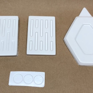 May include: Four white plastic molds for making soap or candles. The molds are in different shapes, including a rectangle, a hexagon, and a long rectangle with three circles.