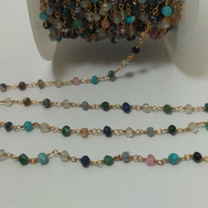 Op de afbeelding: Een gouden ketting met verschillende gekleurde edelsteen kralen, waaronder blauw, groen en wit. De ketting ligt op een witte achtergrond.