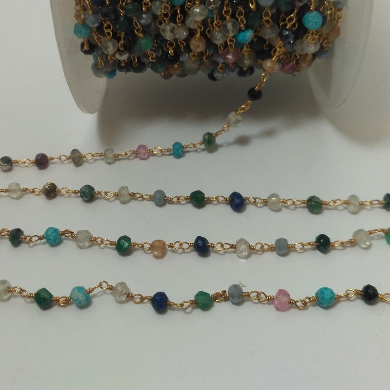 Op de afbeelding: Een gouden ketting met verschillende gekleurde edelsteen kralen, waaronder blauw, groen en wit. De ketting ligt op een witte achtergrond.