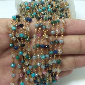 Op de afbeelding: Een veelkleurige edelsteenketting met goudkleurige metalen schakels. De ketting is gemaakt van verschillende edelstenen, waaronder turquoise, lapis lazuli en smaragd.