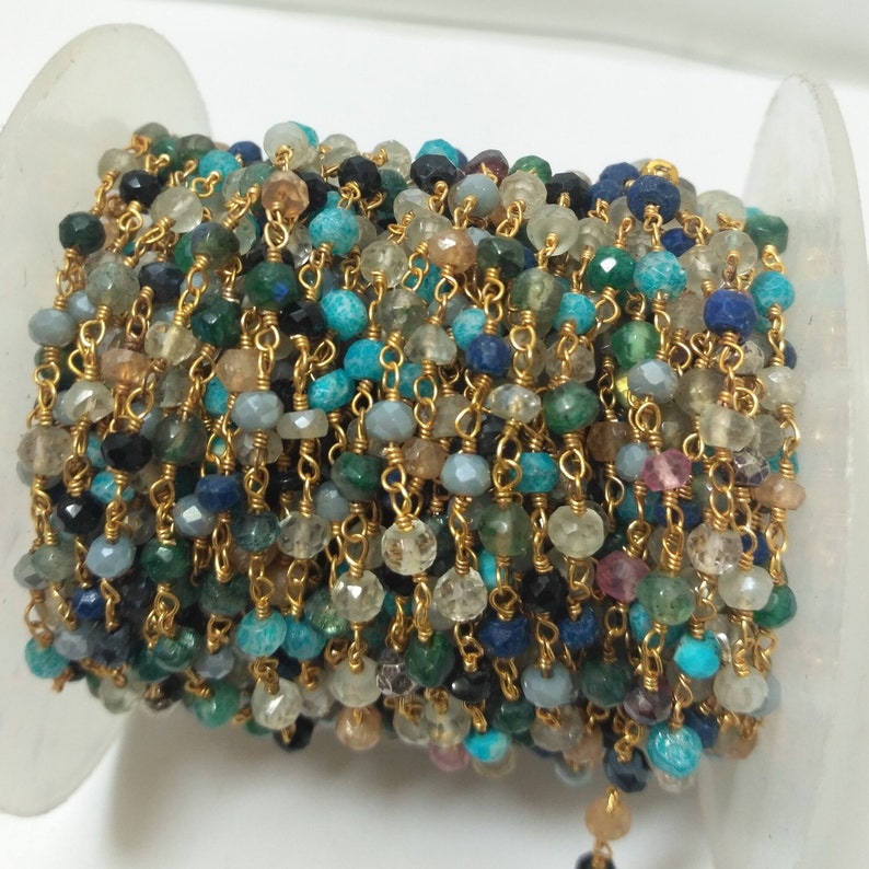 Op de afbeelding: Een veelkleurige edelsteenketting met goudkleurige metalen schakels. De ketting is gemaakt van verschillende edelstenen, waaronder turquoise, blauw, groen en zwart.